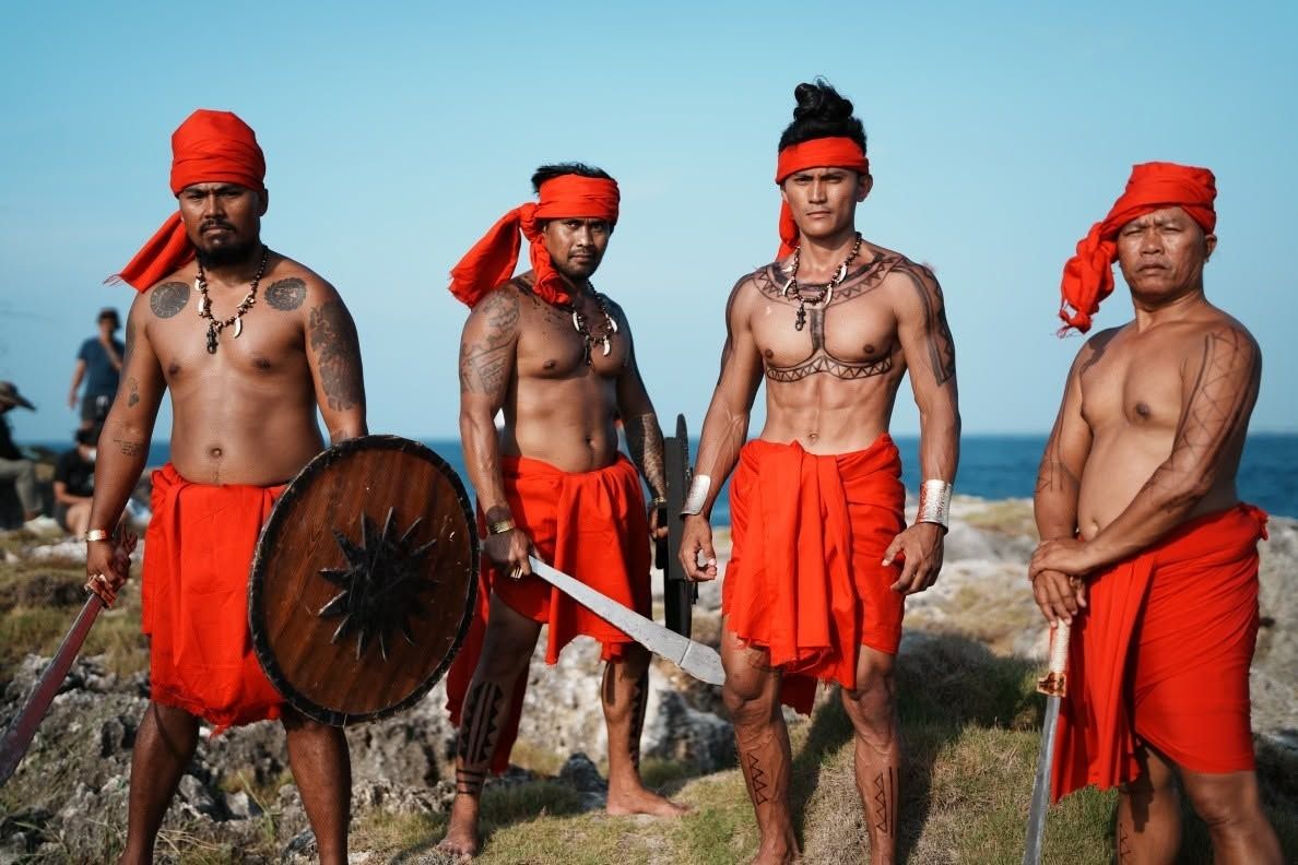 Four warriors in red headcloths and sarongs, with a round shield and a sword, against a blue sky