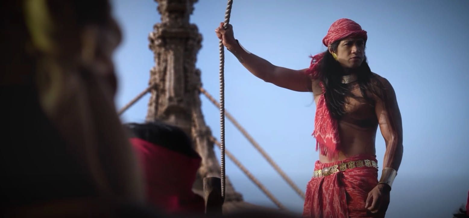 A warrior in a red headcloth and sarong holds a rope in the rigging of a ship