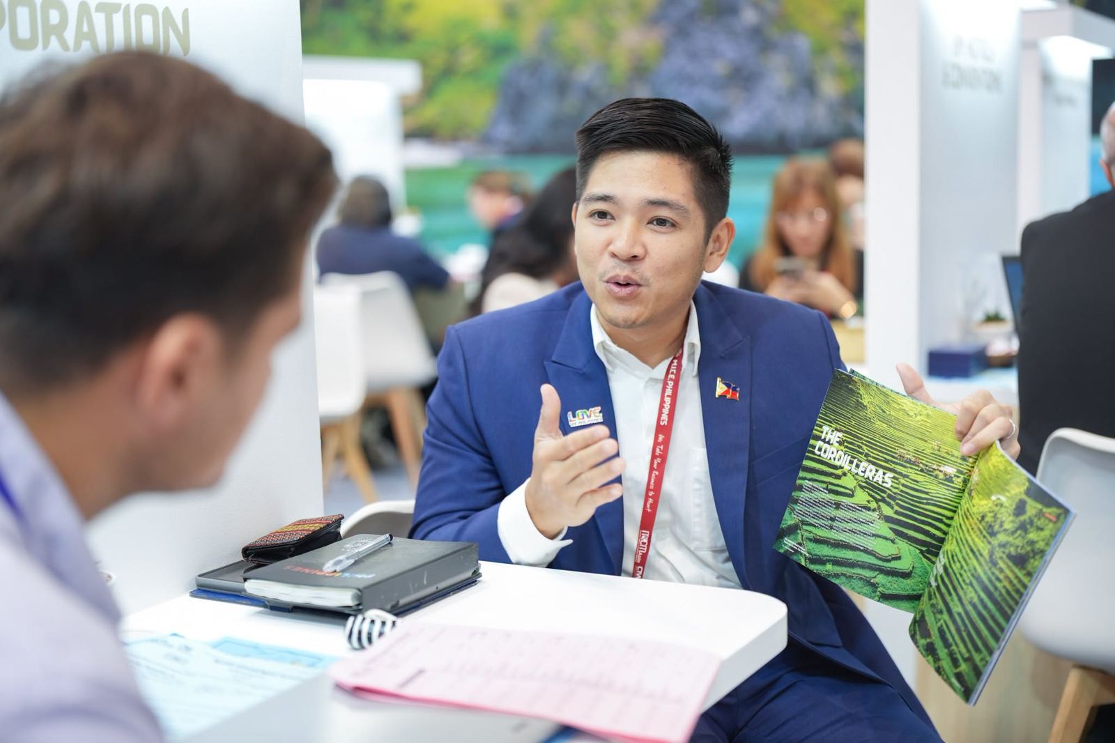 A Philippine exhibitor in a navy suit shows a buyer a brochure on the Cordilleras