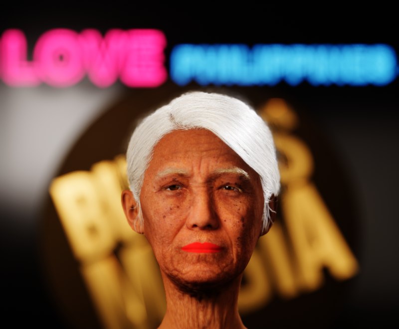 MetaHuman of Apo Whang-Od: white hair, red lipstick, deeply wrinkled skin, in front of a gold Budots Media logo and a neon LOVE PHILIPPINES sign
